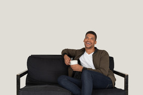 一个微笑的年轻人坐在沙发上，拿着一杯咖啡的前视图