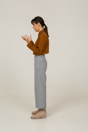 Side view of a questioning young asian female in breeches and blouse raising hands