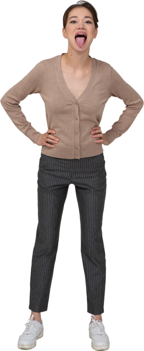 Front view of a young lady in pullover and pants putting hands on hips and showing tongue