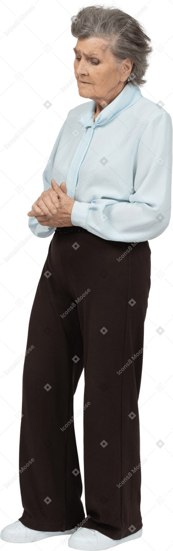 Три четверти вида недовольной пожилой женщины в блузке и брюках, держащей руки вместе и смотрящей вниз