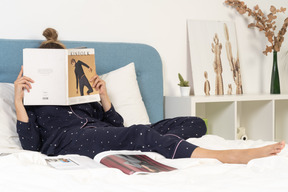 패션 잡지를 읽는 동안 침대에 누워 파자마를 입은 젊은 여성의 전체 길이