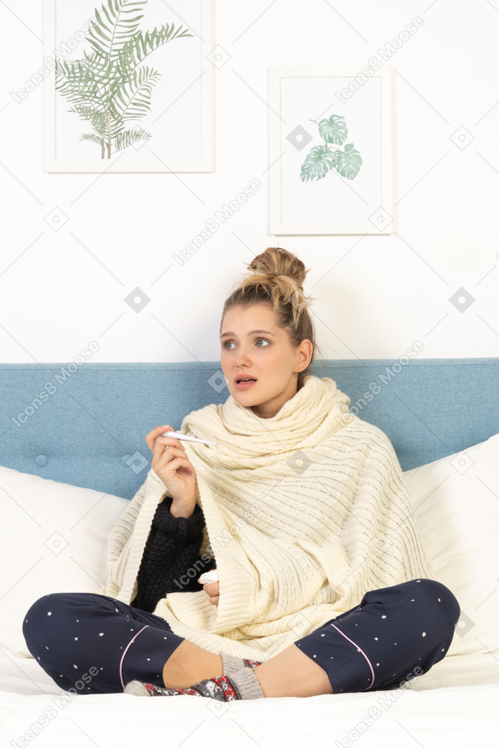 Vista frontale di una giovane donna perplessa avvolta in una coperta bianca seduta a letto con termometro