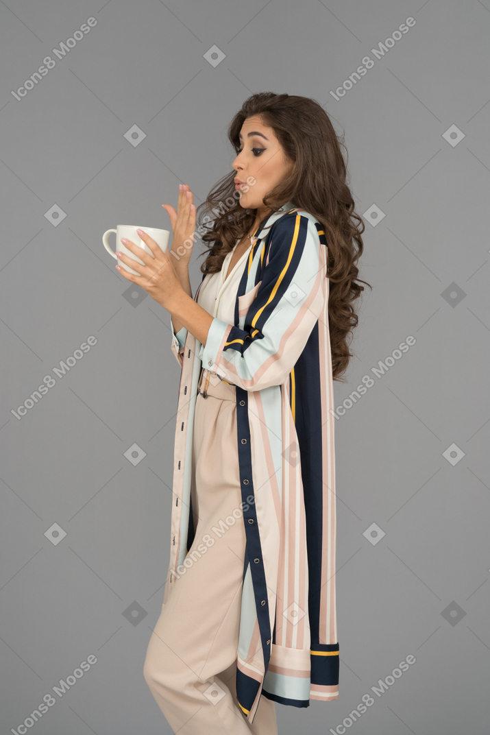 Young woman holding a hot cup of tea and blowing on her right hand