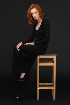 A side view of the cute young woman sitting on the wooden chair and looking to the camera
