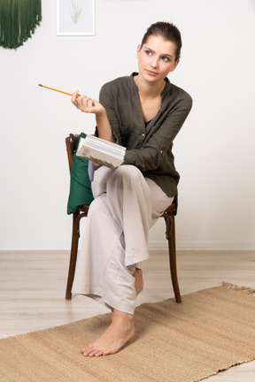 Vorderansicht einer nachdenklichen jungen frau in hauskleidung, die mit bleistift und notizbuch auf einem stuhl sitzt