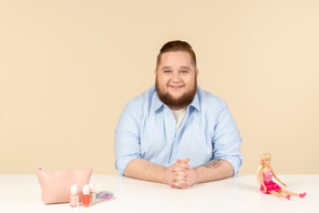 Sonriente tímido joven grande sentado a la mesa y sosteniendo la muñeca barbie