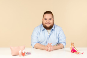 Smiling shy young big man sitting at the table and holding barbie doll