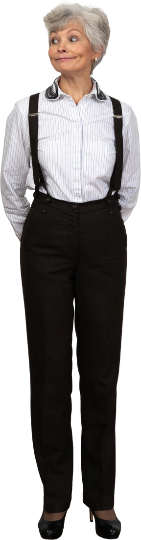 Front view of an old smiling female in office clothes grimacing with her hands behind back looking aside