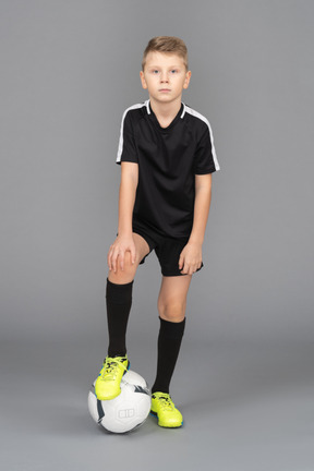 Vista frontal de un niño chico en uniforme de fútbol poniendo su pie en la pelota y mirando a la cámara