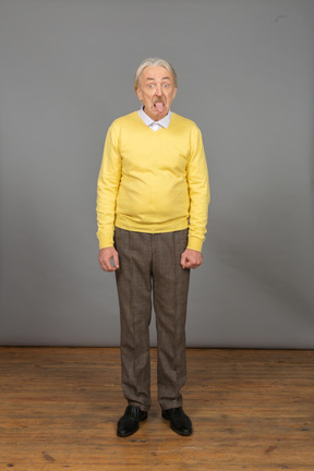 Vista frontal de um velho curioso de blusa amarela, mostrando a língua e olhando para a câmera