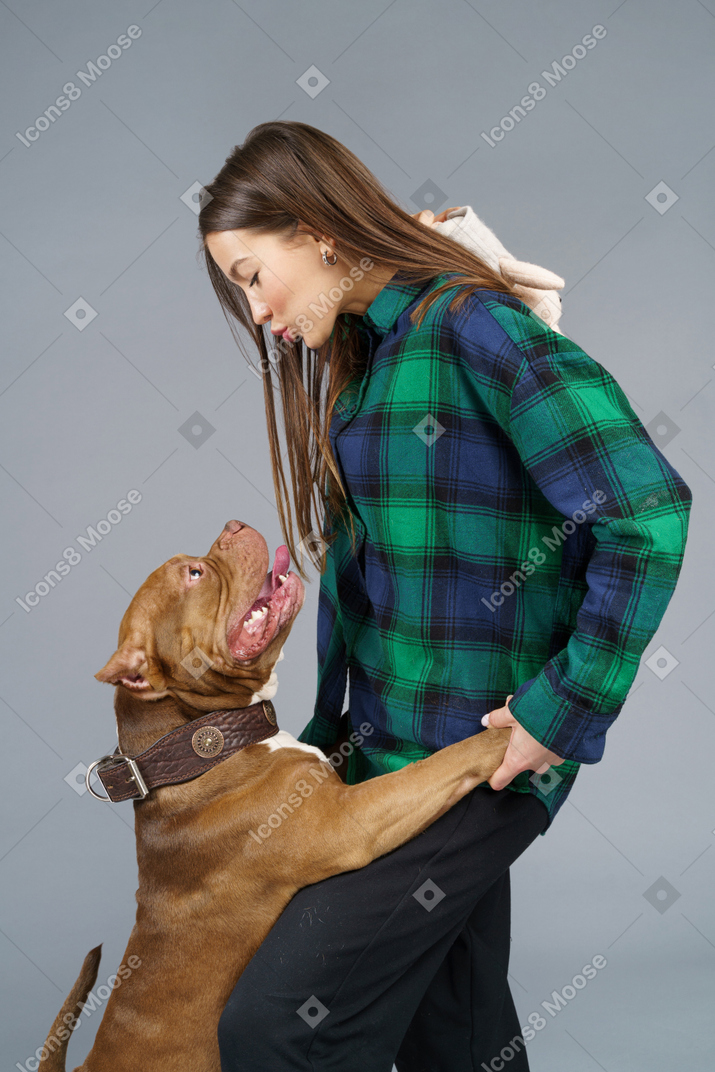 Vista sid de una mujer joven en camisa a cuadros besando a su bulldog marrón