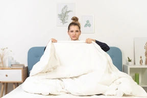 Front view of a grimacing young woman in pajamas hiding behind the blanket staying in bed