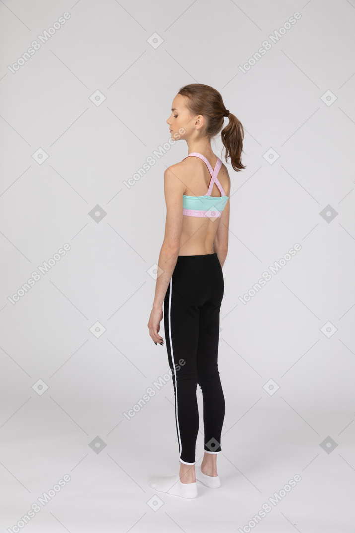 Три четверти сзади девушки-подростка в спортивной одежде, стоящей на месте и смотрящей вниз