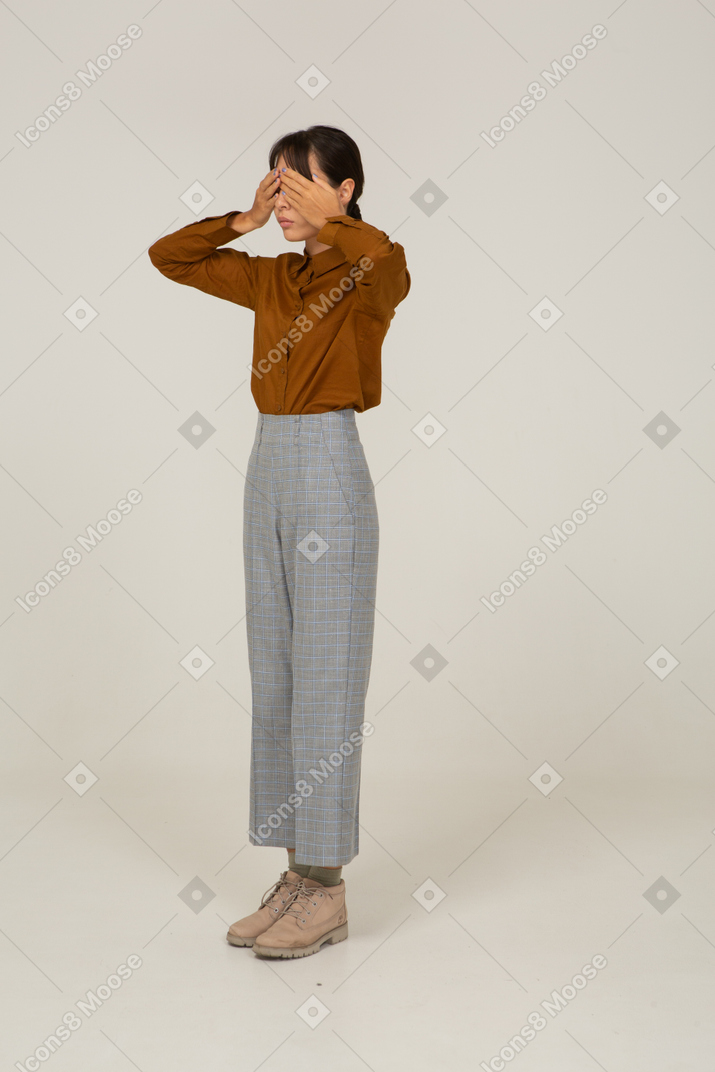 Vista di tre quarti di una giovane donna asiatica in calzoni e camicetta che nasconde gli occhi