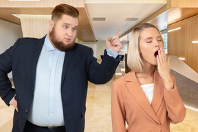 A man in a suit standing and pointing finger behind yawning woman at the office