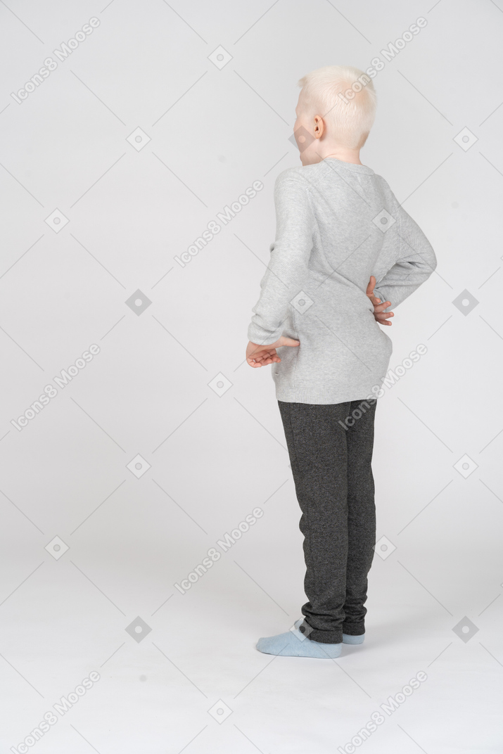 Back three-quarter view of a lonely kid boy in casual clothes putting hand on hip