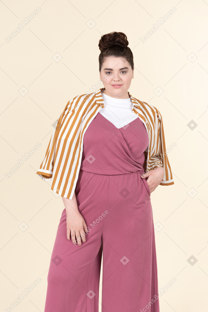 Jeune femme de taille plus dans une combinaison rose et une veste à rayures, posant sur un fond jaune pastel