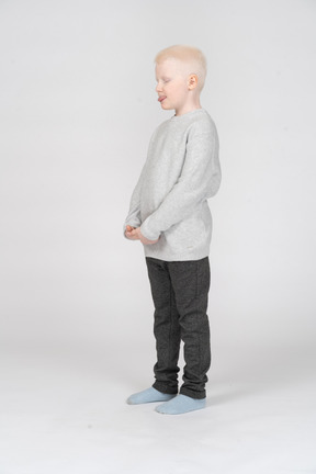 Вид в три четверти мальчика в повседневной одежде, показывающий язык с закрытыми глазами, складывающий руки