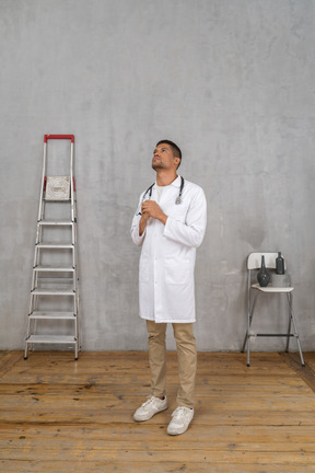 Three-quarter view of a praying young doctor standing in a room with ladder and chair