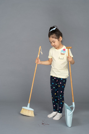 Little girl standing straight while holding broom and dustpan