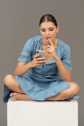 Вид спереди молодой женщины, сидящей на кубе и обменивающейся сообщениями по телефону