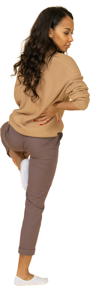 Vista posterior de tres cuartos de una mujer joven de piel oscura que pone la mano en la cadera mientras levanta la pierna