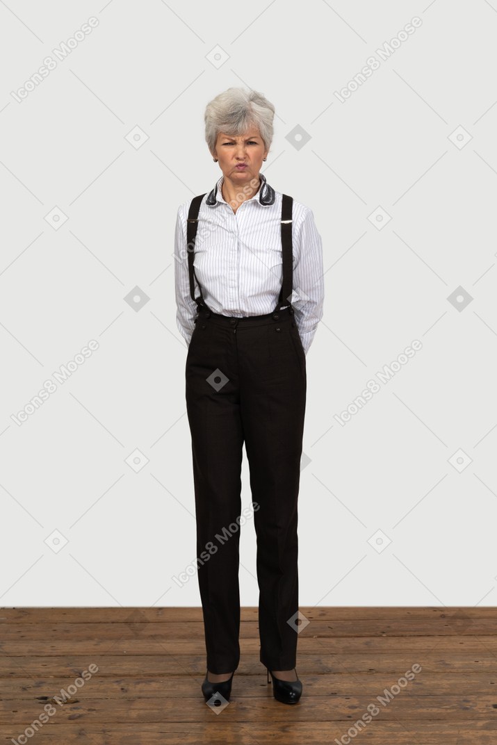 Front view of an old funny female in office clothes grimacing putting hands behind back and knitting brows