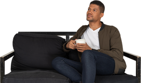 Front view of a young dreaming man sitting on a sofa with a cup of coffee