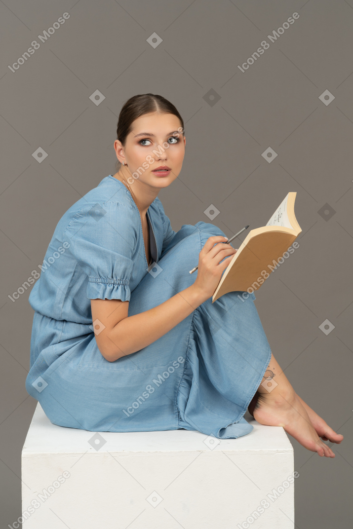 Seitenansicht einer jungen frau, die auf einem würfel sitzt und in ein notizbuch schreibt