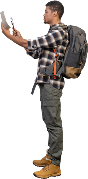 Vista lateral de un turista con una mochila examinando el mapa a través de una lupa