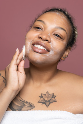 Alegre mujer afro aplicando producto de piel facial con algodón