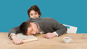 Boy standing behind young man sleeping on table