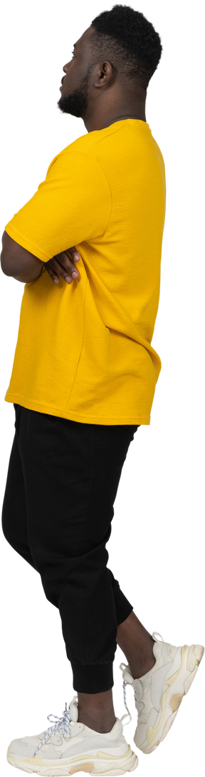 Vista laterale di un giovane uomo sospettoso dalla pelle scura con una maglietta gialla che incrocia le braccia
