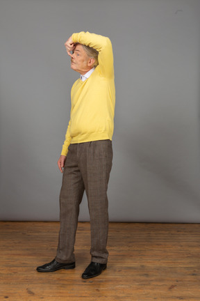 Трехчетвертный вид старого забавного человека в желтом пуловере, касающегося носа и смотрящего в сторону