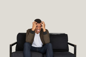 Mad looking young man sitting on the sofa and touching his head with hands