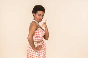 Young black short-haired woman in a checkered top and a skirt, posing against a plain peachy background