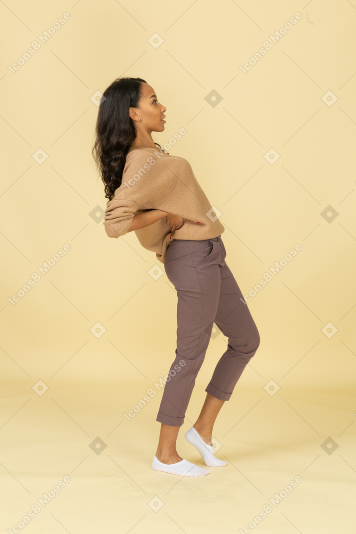 Vista lateral de una mujer joven de piel oscura poniendo las manos en las caderas y recostándose