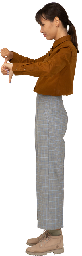 Vista lateral de uma jovem mulher asiática de calça e blusa com os polegares para baixo