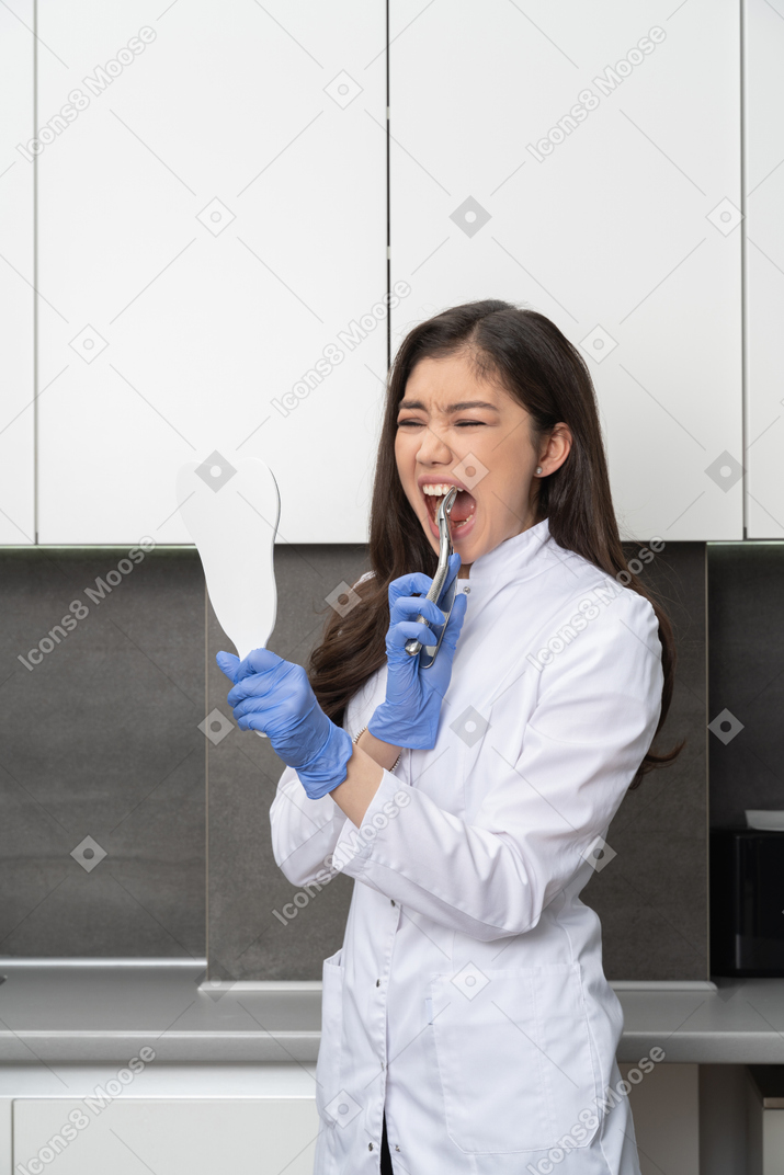Vista de três quartos de uma médica assustada, olhando-se no espelho e tocando os dentes com um instrumento dentário