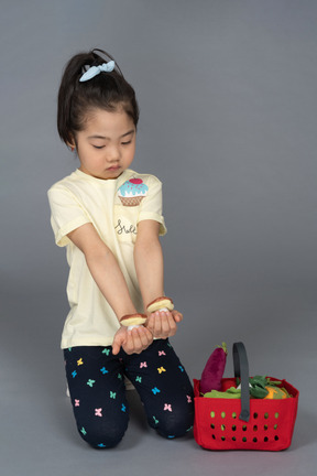 Little girl holding two mushrooms while sitting beside a shopping basket