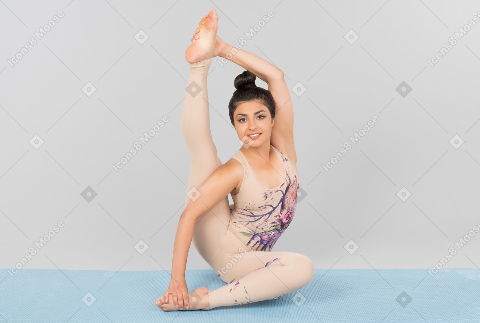 Joven gimnasta india sentada sobre una estera de yoga con una pierna arriba y tocando su dedo del pie con una mano