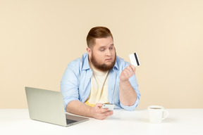 Young overweight man sitting at the table, holding phone and looking at bank card