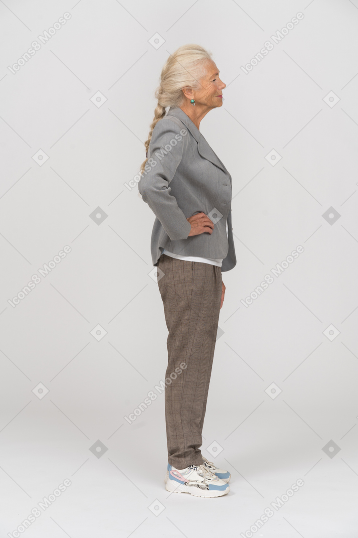 Вид сбоку пожилой женщины в костюме, позирующей с руками на бедрах
