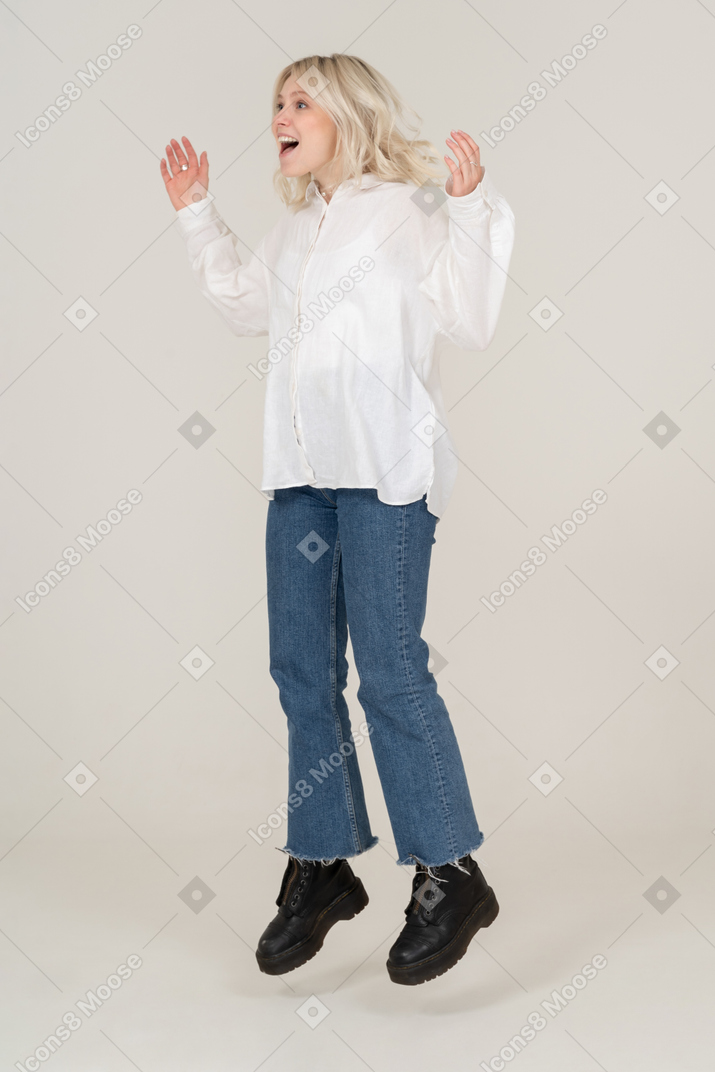 Vista di tre quarti di una giovane donna sorpresa in abiti casual, saltando e alzando le mani