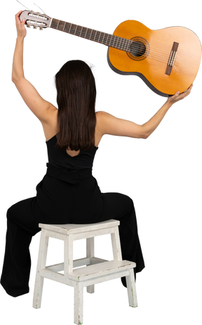 Vista posteriore di una giovane donna in abito nero che tiene la chitarra sopra la testa e seduto su uno sgabello