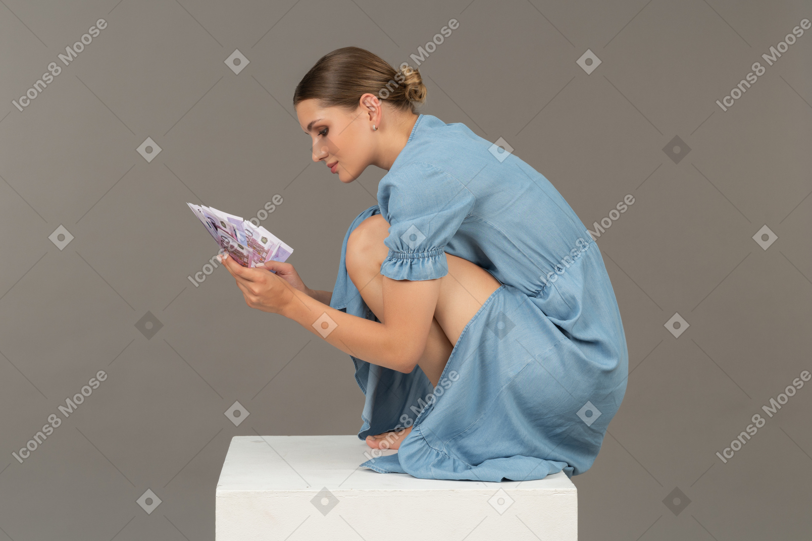 Side view of young woman sitting on cube with bunch of banknotes