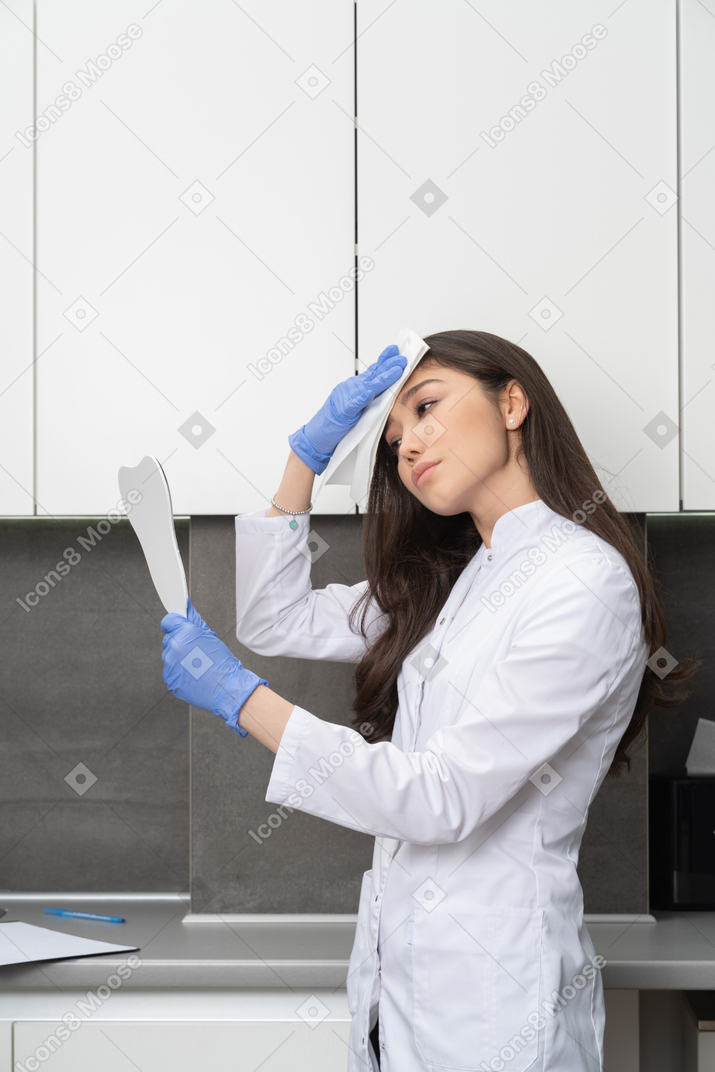 Side view of a female doctor looking in the mirror and wiping her forehead