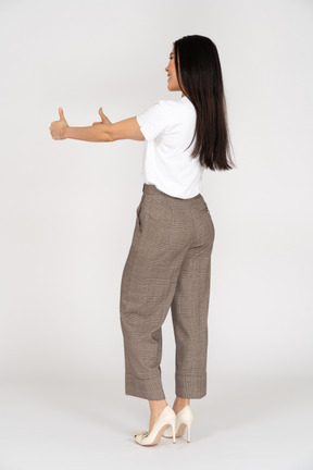 Vista traseira a três quartos de uma jovem sorridente de calça e camiseta mostrando os polegares para cima
