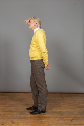Vista laterale di un vecchio uomo divertente in pullover giallo che tocca il naso e che osserva da parte