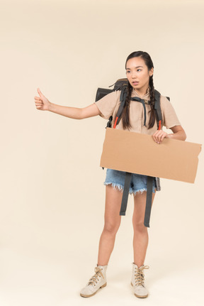 Young female hitchhiker holding paper card and showing thumb up